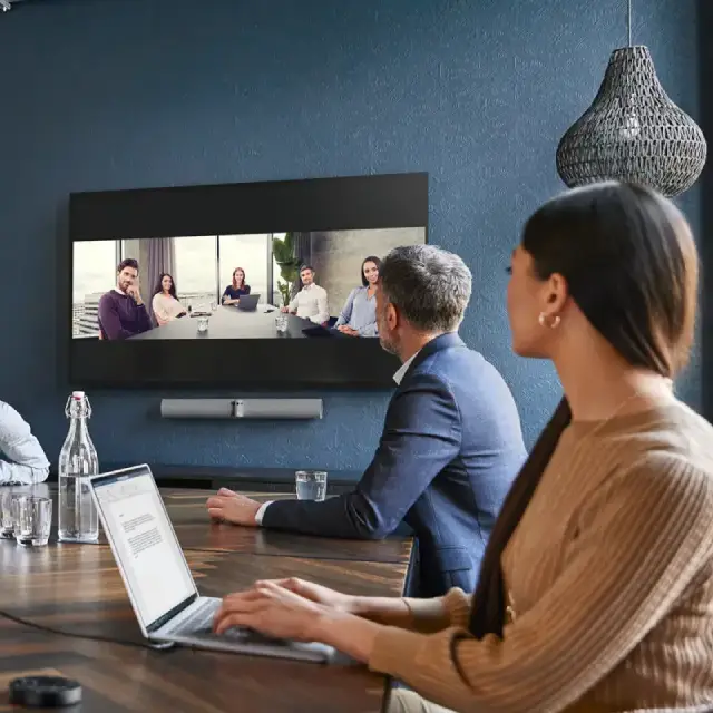 Dos personas en una oficina mirando una pantalla de TV y computador portátil en conferencia con otro equipo de trabajo.