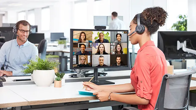 Una mujer en una reunión virtual con nueve personas en el monitor del computador. En frente hombre con diadema dando soporte.