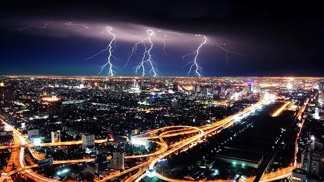 Ciudad en la noche con todas las luces iluminando carreteras, edificios y casas. Al fondo rayos en el cielo.